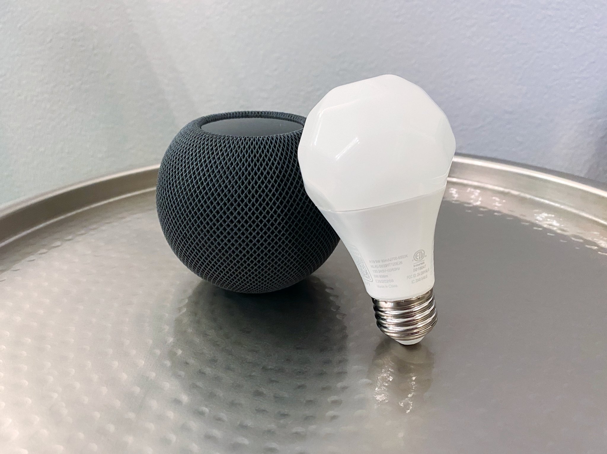 Nanoleaf Esssentials A19 Light Bulb leaning on a Space Gray HomePod mini