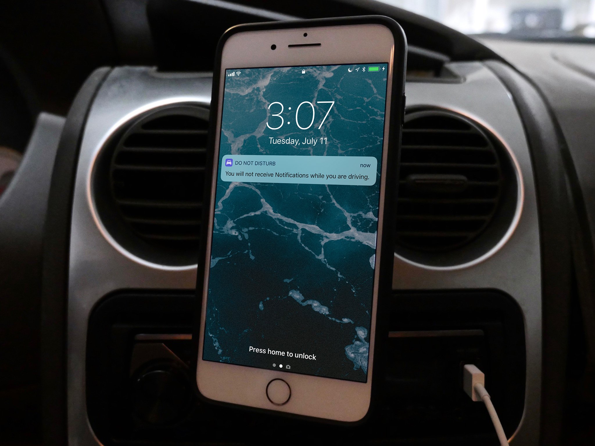 An iPhone is shown attached to a car mount. A notification shows Do Not Disturb While Driving is activated.