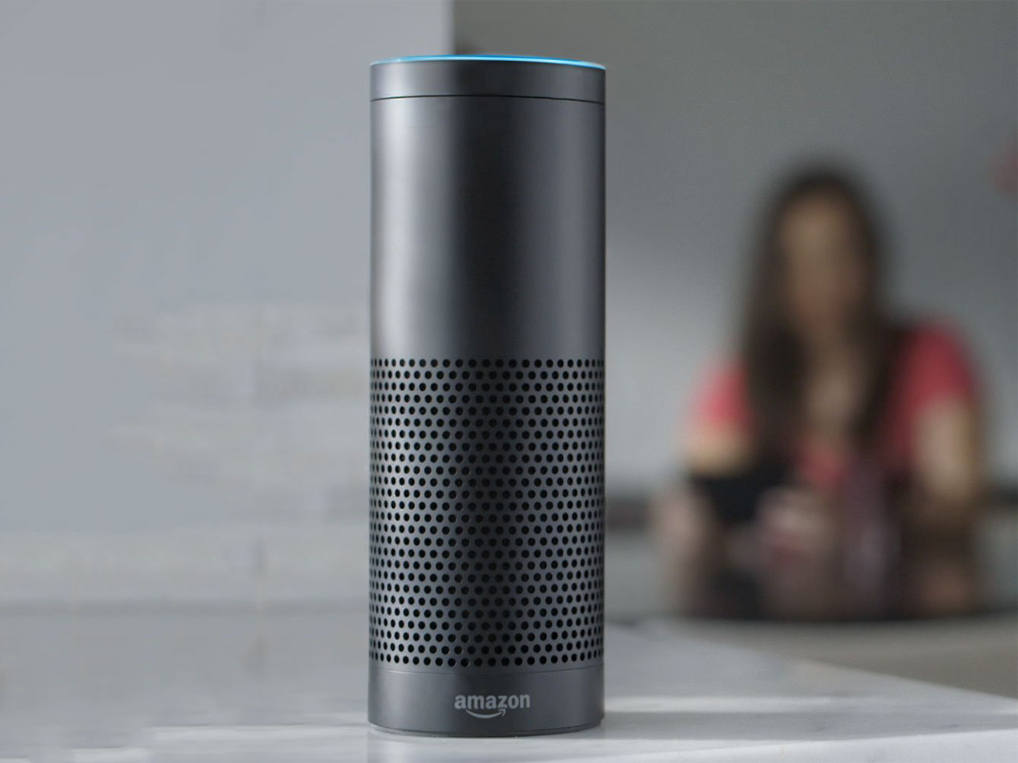 An Amazon Echo is shown sitting on a table. A person sits nearby.