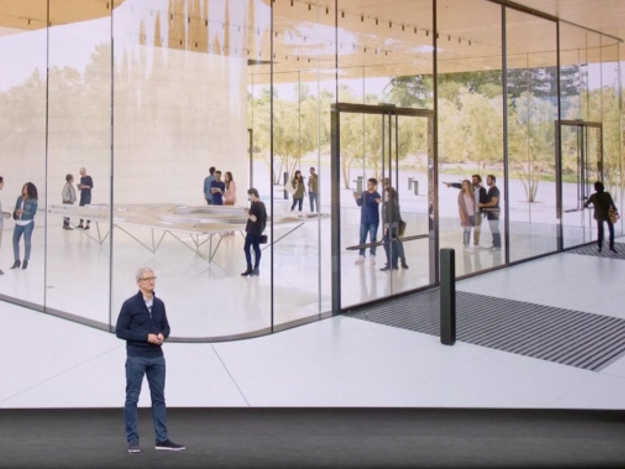 Tim Cook stands on stage in front of a photograph of Apple Park&#39;s Visitor Center
